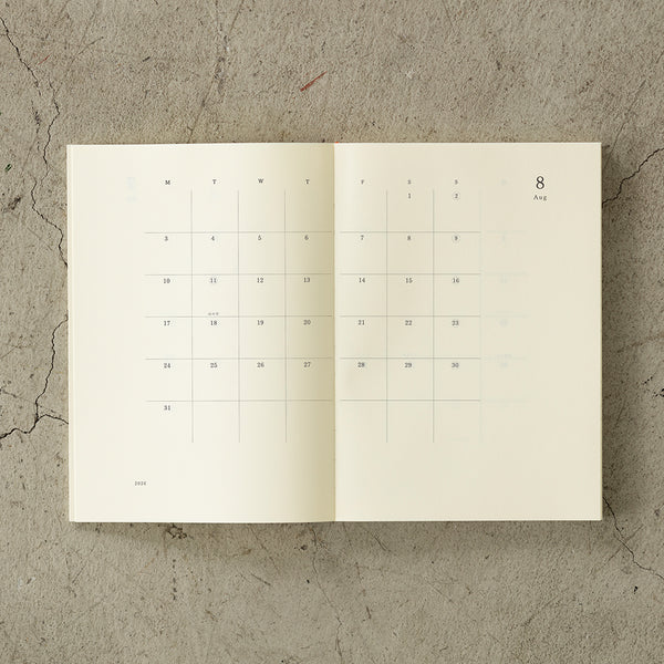 Load image into Gallery viewer, A5 MD Notebook Diary 2026 with a minimalist cream-toned glassine paper cover, thread-stitched binding, and a white bookmark string. The open spread displays a block-style monthly calendar on one side and lined memo pages on the other, printed on smooth MD PAPER designed for a premium writing experience.