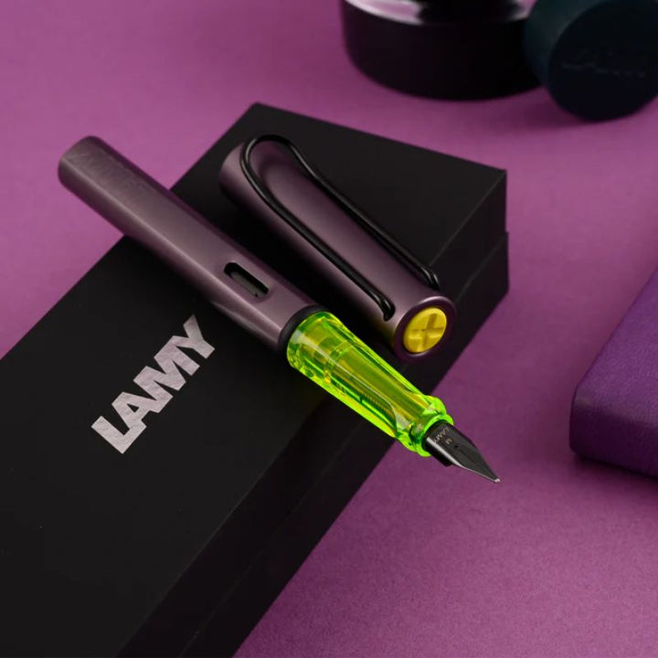 LAMY AL-star fountain pen in metallic aubergine with a translucent neon yellow grip, black steel nib, and metal clip. Includes a blue ink cartridge.