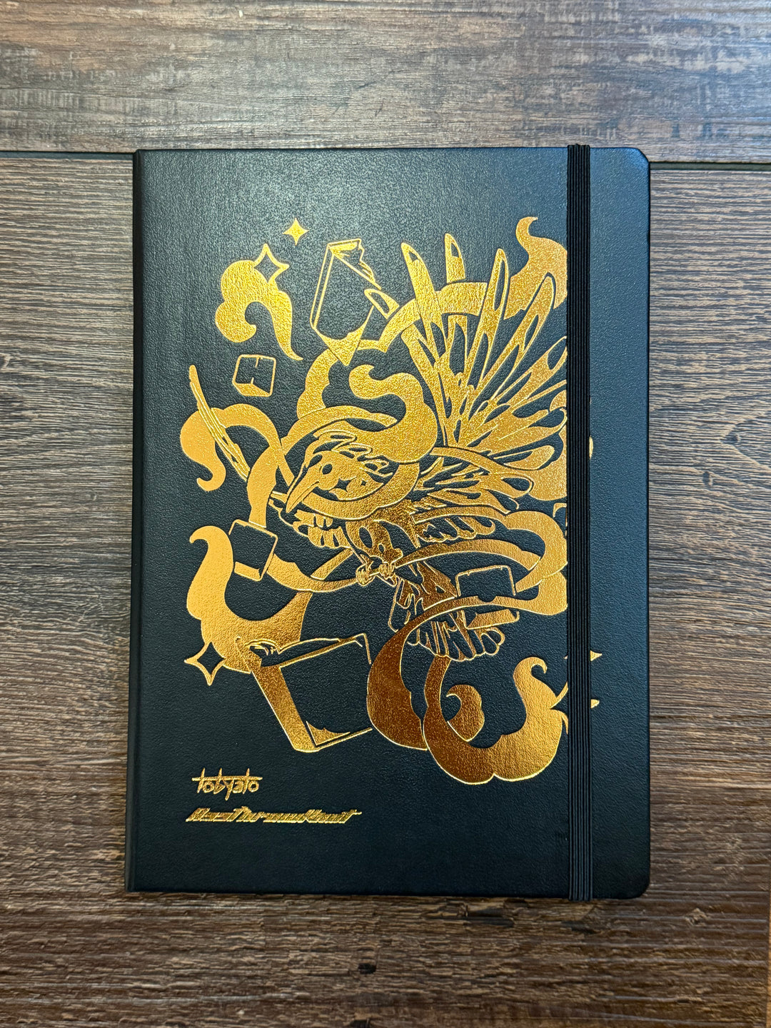 Leuchtturm1917 Tobyato Edition A5 Medium Notebook in black with ruled pages, featuring a cover design of a raven surrounded by books and typewriter keys, symbolizing creativity and knowledge.