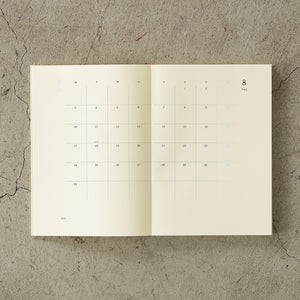 A5 MD Notebook Diary 2026 with a minimalist cream-toned glassine paper cover, thread-stitched binding, and a white bookmark string. The open spread displays a block-style monthly calendar on one side and lined memo pages on the other, printed on smooth MD PAPER designed for a premium writing experience.