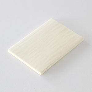 A5 MD Notebook Diary 2026 with a minimalist cream-toned glassine paper cover, thread-stitched binding, and a white bookmark string. The open spread displays a block-style monthly calendar on one side and lined memo pages on the other, printed on smooth MD PAPER designed for a premium writing experience.