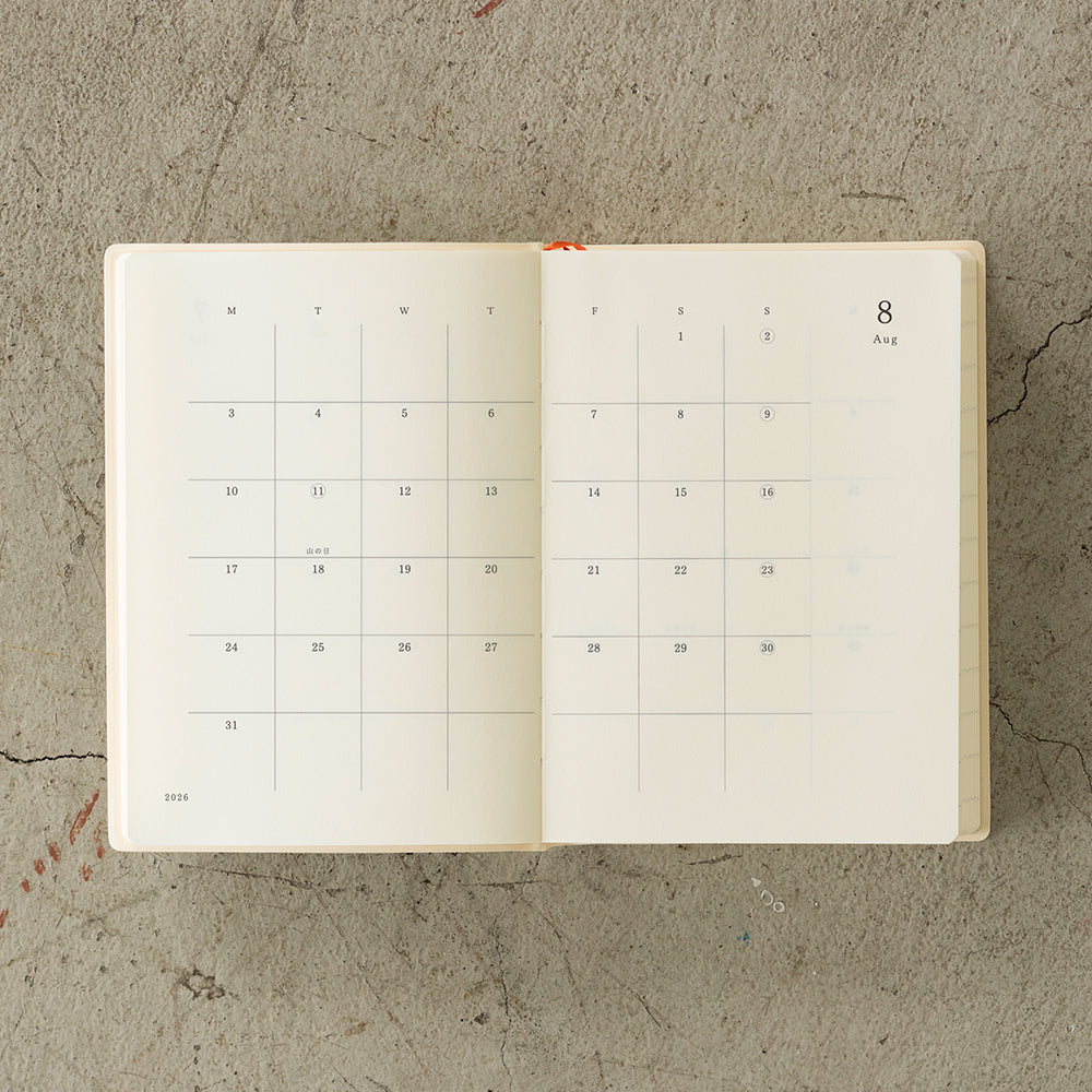 MD Notebook Diary (A6) 1 Day 1 Page 2026 with a minimalist PVC cover, featuring a year stamp on the spine, two bookmark strings, and monthly index tabs. The diary is open to reveal its premium MD PAPER with a clean, spacious layout for daily entries.